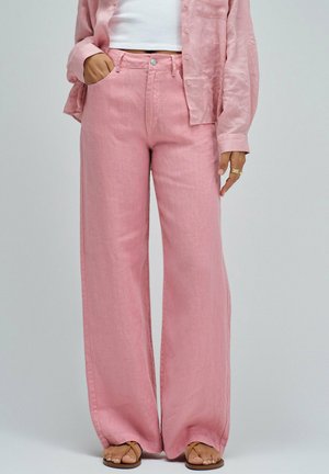 Mujer con pantalones anchos rosas, camisa rosa a juego, camiseta blanca y sandalias marrones, mano en el bolsillo, de pie frente a un fondo gris.