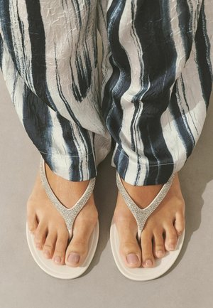 Feet wearing white flip-flops with rhinestone straps and loose pants featuring black and white vertical abstract stripes.