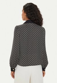 Donna con lunghi capelli ricci che indossa una blusa a righe bianche e nere con motivi a "M" e catena ripetuti, abbinata a pantaloni bianchi, di spalle.