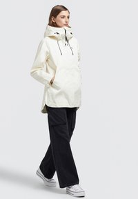 Jachetă anorak ușoară, de culoare crem, cu glugă ajustabilă, buzunare laterale și o croială lejeră, asortată cu pantaloni cargo negri și teniși albi.