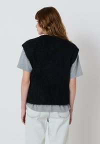 Moteris su banguotais rudais plaukais, vilkinti juodą purią be rankovių liemenę ant pilkos trumpomis rankovėmis marškinėlių ir šviesiai mėlynų džinsų, atsisukusi nugara prie baltos sienos.