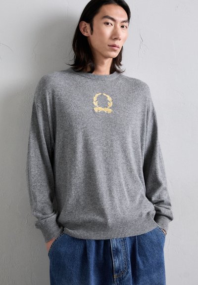 Grau gestrickter Pullover mit geripptem Saum, der ein goldenes besticktes Kranzlogo auf der Brust trägt. Getragen mit lockeren blauen Jeanshosen.