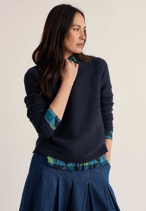 Kvinde med langt mørkt hår iført en marineblå sweater over en blå ternet skjorte og en denimnederdel, kigger til siden foran en ensfarvet baggrund.