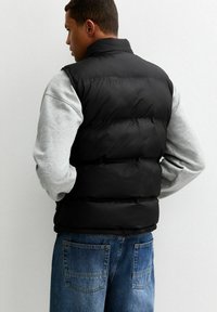 Czarna kamizelka puchowa z wysokim kołnierzem, pikowanym wzorem i elastycznym dołem. Noszona na szarej koszuli z długim rękawem, połączona z niebieskimi dżinsami.