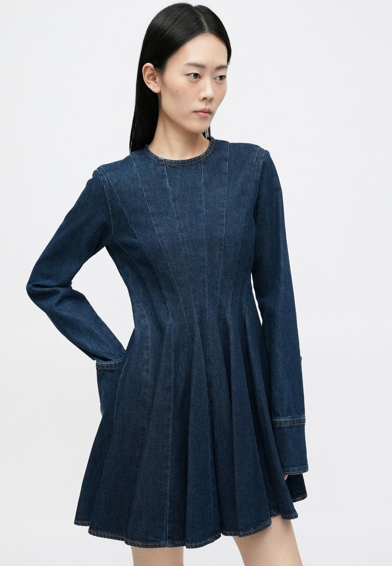 Robe en denim à manches longues, corsage ajusté et jupe évasée. Couleur bleu foncé avec des détails de coutures et un col rond.