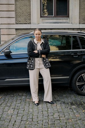 Jeune femme en tenue beige et veste noire brodée, bras croisés, à côté d’un SUV noir dans une rue pavée.