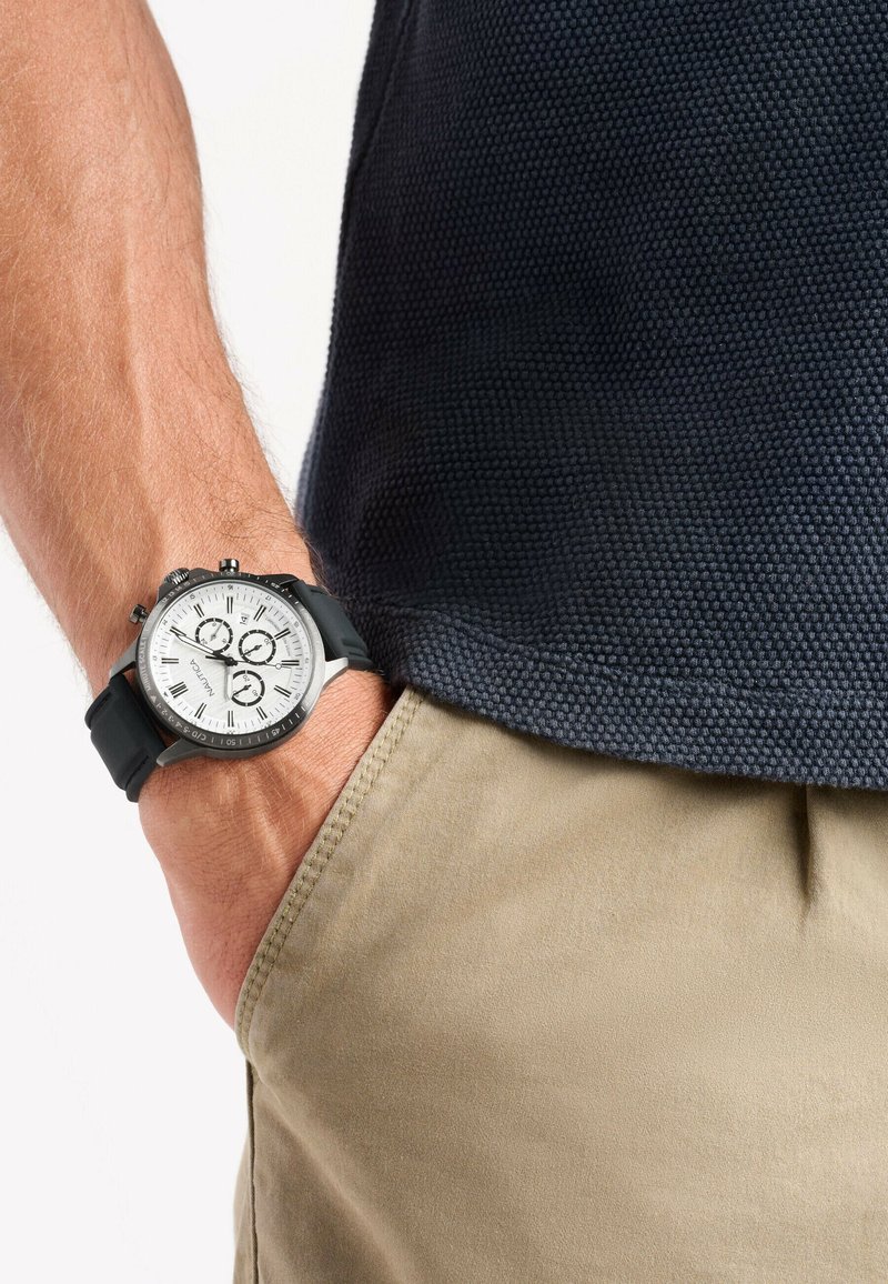 Reloj cronógrafo negro con esfera blanca, tres subesferas y correa de goma, combinado con una camisa texturizada azul marino y pantalones cortos beige.