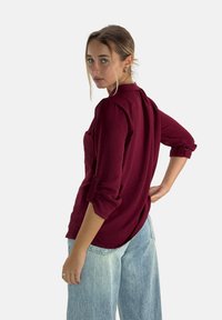 Camicia bordeaux a maniche lunghe con polsini arrotolati, tessuto testurizzato, orlo curvo e tasca frontale, abbinata a jeans a gamba larga in un leggero blu.
