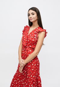 Rochie roșie cu flori, cu mâneci scurte cu volane, decolteu în V și cordon în talie. Materialul are flori mici albe și roz pe un fundal texturat.