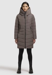Langer, gesteppter Mantel in sanftem Taupe mit hohem Kragen, diagonaler Steppung, Frontreißverschluss und Seitentaschen. Getragen über schwarzen Leggings und Knöchelstiefeln.