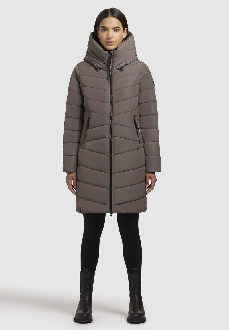 Langer, gesteppter Mantel in sanftem Taupe mit hohem Kragen, diagonaler Steppung, Frontreißverschluss und Seitentaschen. Getragen über schwarzen Leggings und Knöchelstiefeln.