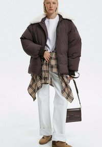 Brązowa kurtka puchowa z kołnierzem z sztucznego futra, biały t-shirt, koszula w kratę przewiązana w pasie, białe szerokie spodnie, brązowa torba i beżowe sneakersy.