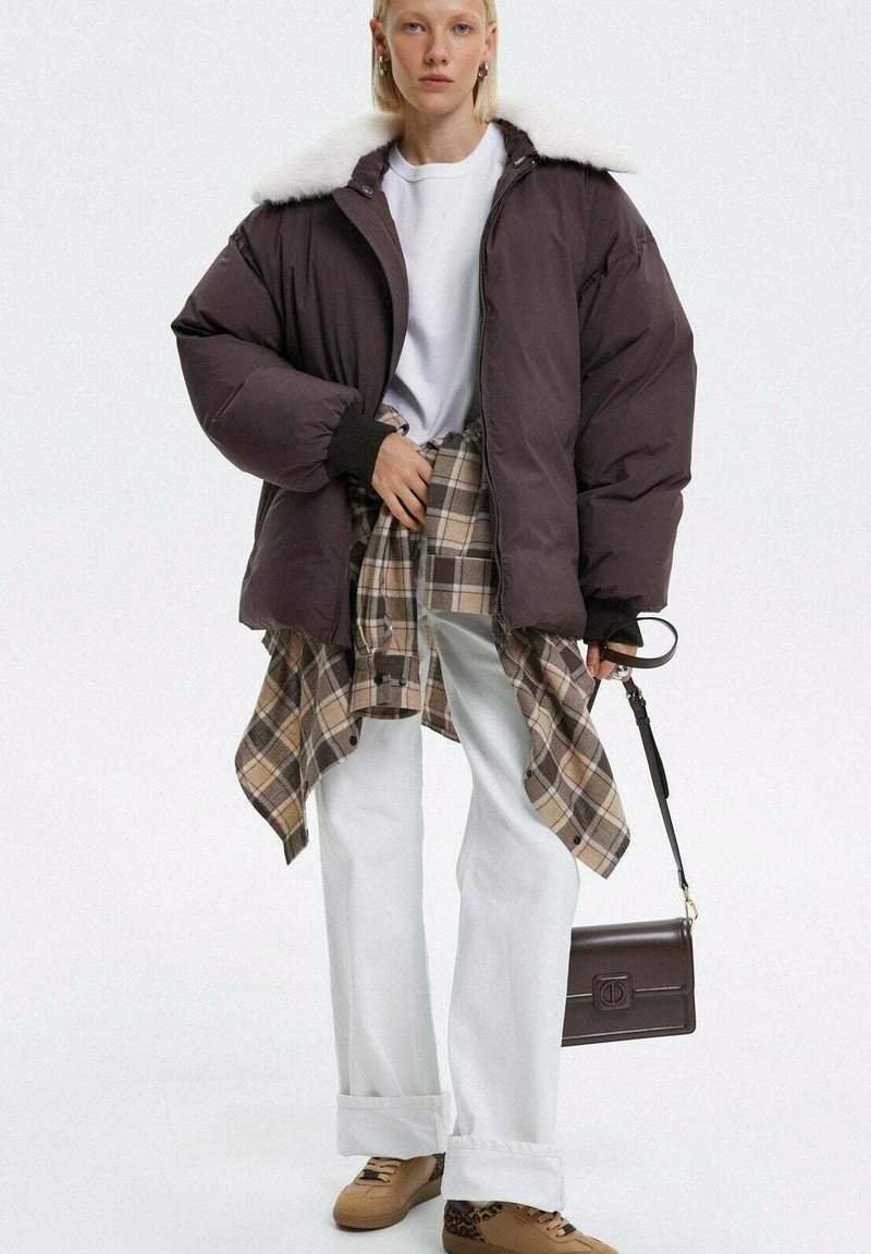 Brązowa kurtka puchowa z kołnierzem z sztucznego futra, biały t-shirt, koszula w kratę przewiązana w pasie, białe szerokie spodnie, brązowa torba i beżowe sneakersy.