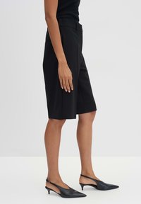 Schwarze, maßgeschneiderte Shorts mit geradem Schnitt, in mittlerer Oberschenkel-Länge, mit Falten und seitlichen Taschen. Kombiniert mit schwarzen Slingback-Absätzen.