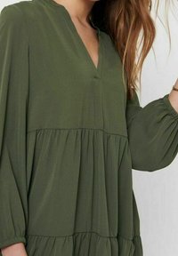 Olivengrünes Kleid mit V-Ausschnitt, plissiertem Oberteil und gestuftem Design. Lange Puffärmel mit elastischen Bündchen. Weicher, strukturierter Stoff.