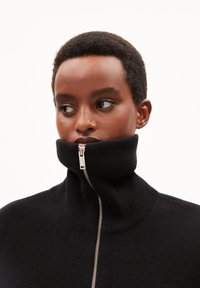 Suéter negro de punto con cuello alto y cremallera, que presenta una cremallera plateada. Textura suave con detalles acanalados en el cuello para añadir contraste.