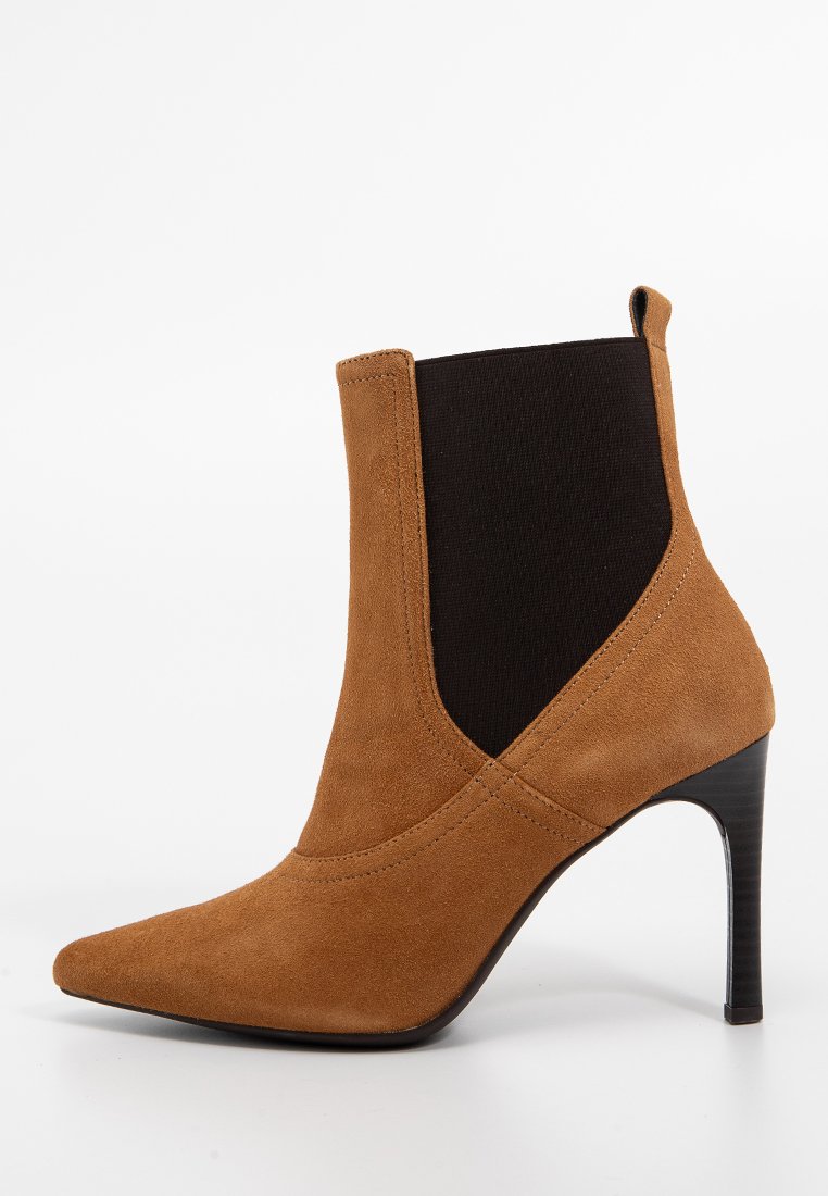 Stivale alla caviglia in suede marrone con punta affusolata e un pannello elastico nero. Presenta un tacco alto e sottile e dettagli cuciti ai lati.