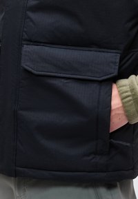 Χέρι στην τσέπη ενός μαύρου επενδυμένου μπουφάν με τσέπη με καπάκι, φορεμένο πάνω από ένα πράσινο μανίκι φλις και γκρίζο παντελόνι.