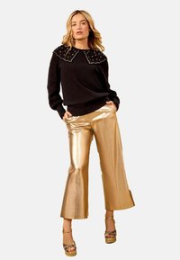 Pulover negru cu guler în care sunt incrustate ținte, purtat cu pantaloni largi cu un aspect lucios de culoare aurie. Modelul poartă sandale cu platformă și curea texturată.