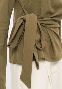 Μακρυμάνικη μπλούζα σε ελιά πράσινο χρώμα δεμένη σε μεγάλο φιόγκο στη μέση, συνδυασμένη με λευκό παντελόνι.