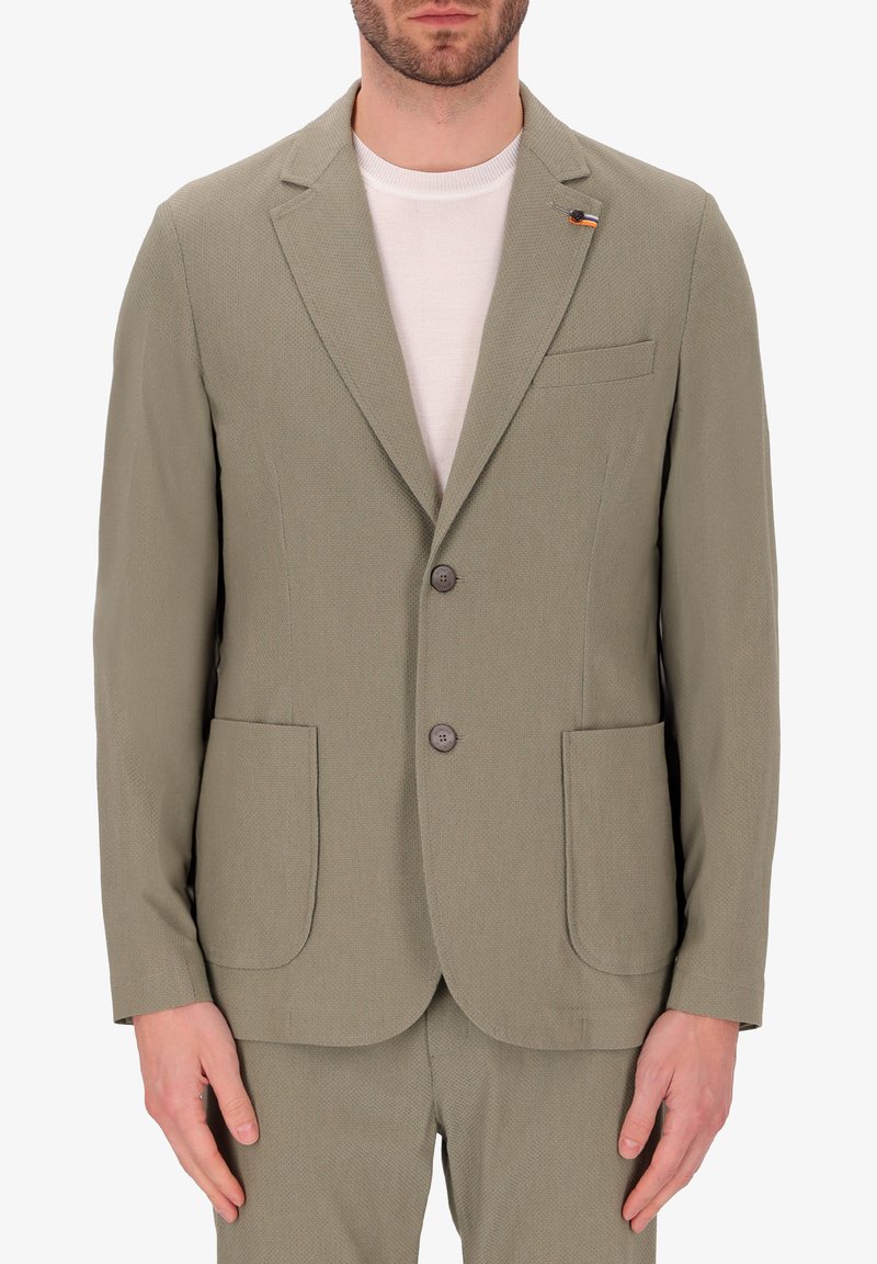 Blazer beige con texture, dotato di due tasche frontali, due bottoni e bavero a punta. Presenta una piccola spilla a forma di bandiera sul lato sinistro.