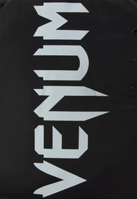 Sac de sport noir arborant un lettrage "VENUM" en blanc audacieux, avec une texture mate et des éléments de design angulaires. Comprend des fermetures à pression.