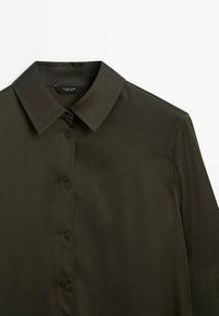 Chemise vert olive foncé à boutons, col classique et manches longues, présentée sur fond blanc.
