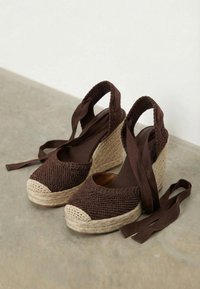 Bruine gevlochten sleehakken espadrilles met beige gevlochten platformzolen en enkelbandjes, geplaatst op een grijs oppervlak tegen een witte achtergrond.