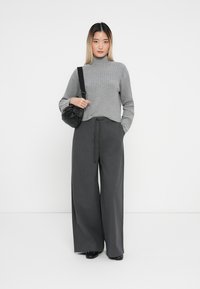 Grau gerippter Rollkragenpullover kombiniert mit hoch taillierten, weiten anthrazitfarbenen Hosen. Das Modell trägt eine kleine schwarze Umhängetasche.