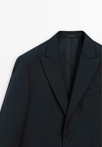 Granatowa marynarka z szpiczastymi klapami, zapinana na jeden guzik, z kieszenią na piersi. Gładka tekstura i dopasowany krój, z minimalnymi detalami ściegów.