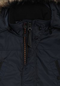 Chaqueta de plumas azul marino con capucha forrada de piel, cuello acanalado negro, cremallera frontal con acento naranja y botones a presión en los bolsillos.