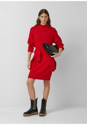 Rode gebreide jurk met een hoge hals, geribbelde manchetten en een strikceintuur. Gecombineerd met zwarte enkellaarsjes met een robuuste zool en textuur zijpanelen.