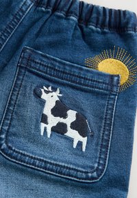 Shorts in denim con cintura elastica; presenta una mucca ricamata blu e bianca sulla tasca posteriore, accanto a un design di sole giallo.