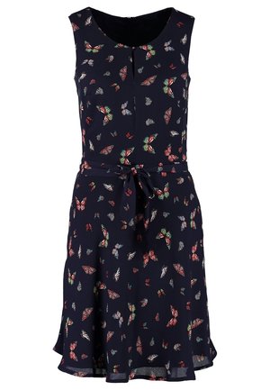 Femme portant une robe sans manches bleu marine ornée d'un imprimé papillons colorés, des collants noirs, des mocassins en cuir noir et un collier noir en perles.