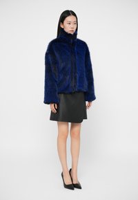 Γυναίκα με μπλε ψεύτικη γούνα, μαύρη δερμάτινη μίνι φούστα και μαύρες ψηλές γόβες, στέκεται μπροστά από ένα απλό ανοιχτό φόντο.