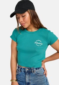Aqua groene cropped t-shirt met ribbeltextuur, voorzien van een witte "Billabong" logo. Gecombineerd met hoge spijker shorts en een zwarte pet.