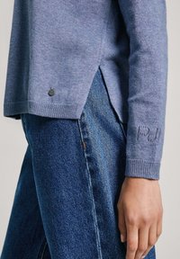 Pull tricoté bleu clair avec poignets côtelés, fentes latérales et un petit logo noir en accent. Associé à un jean en denim avec une couture visible.