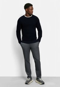 Marineblauer Strickpullover mit kontrastierendem weißem Kragen, kombiniert mit grauen, strukturierten Hosen und grauen Turnschuhen, präsentiert von einer stehenden männlichen Figur.