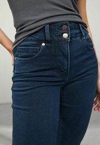 Los jeans de mezclilla azul oscuro presentan una cintura alta, un cierre delantero con botón y un estilo clásico de cinco bolsillos con discretos acentos de costura.
