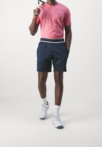 Růžová košile s krátkým rukávem, námořnické sportovní šortky s bílým pasem, bílé tenisky, držící tennisovou raketu. Hladká tkanina, uvolněný střih.