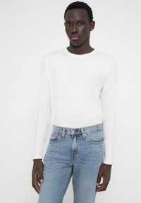 Λευκό βαμβακερό πουκάμισο με μακριά μανίκια και στρογγυλή λαιμόκοψη, συνδυασμένο με ανοιχτό μπλε τζιν παντελόνι. Λείος υφασμάτινος τόνος, κλασική εφαρμογή και χωρίς ορατά σχέδια.