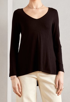 Personne portant un haut noir à manches longues avec un col en V, un pantalon couleur crème, un collier argenté et plusieurs bracelets argentés au poignet gauche.