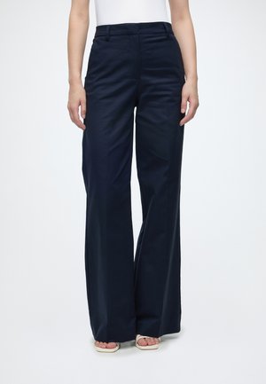 Femme portant un pantalon large taille haute bleu marine avec des poches devant et un haut blanc, debout sur un fond blanc uni.
