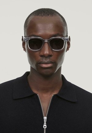 Klare Sonnenbrille mit einem transparenten Gestell, dunklen Gläsern und quadratischer Form. Getragen mit einem schwarzen Polohemd mit Reißverschluss.