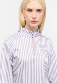 Blond kvinde med blå øjne iført en hvid og grå vertikalt stribet bluse med en knaplukket flæsekant og nøglehulsudskæring.