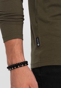 Olivgrünes Langarmhemd mit seitlichem Etikett und glatter Textur. Schwarze Perlenarmband mit metallischen Akzenten am Handgelenk.