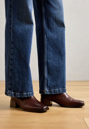 Stivaletti marroni alla caviglia con punta quadrata, tacco basso a blocco e texture in pelle liscia, abbinati a jeans a gamba larga azzurri.