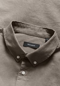 Braune Button-up-Hemd mit klassischem Kragen, weißen Knöpfen und einer Stoffstruktur, die weich und leicht knittrig erscheint. Markenlabel sichtbar.