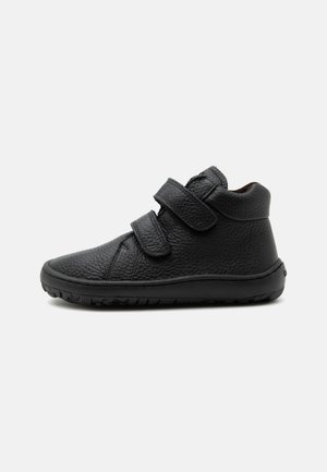 Chaussure montante en cuir noir texturé avec deux bandes auto-agrippantes réglables et une semelle en caoutchouc souple, présentée de profil sur fond blanc.