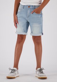 Pantalones cortos de mezclilla azul claro con dobladillos enrollados, que presentan bolsillos frontales y un cierre de botón marrón. Combinados con zapatillas blancas y grises.